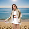 A woman wearing a beige oversized microfiber quick-dry beach towel stands on a sandy beach with the ocean in the background.