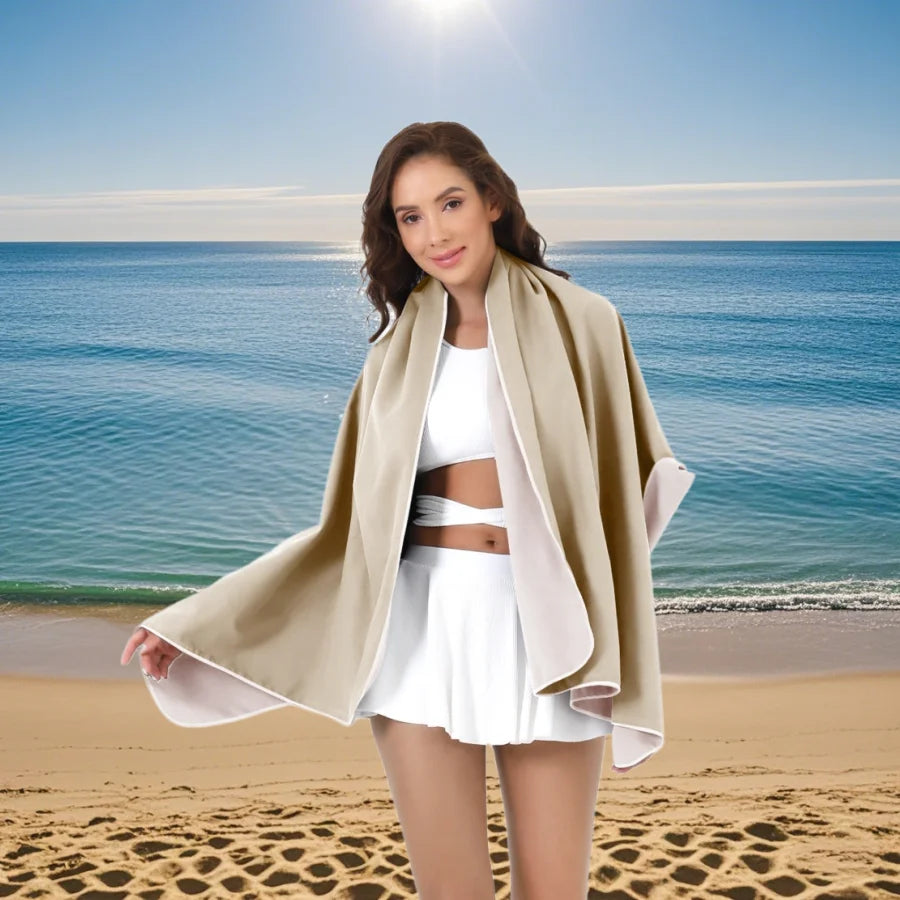 A woman wearing a beige oversized microfiber quick-dry beach towel stands on a sandy beach with the ocean in the background.