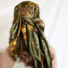 A close-up of a satin square scarf in olive green and gold floral print tied around a person's hair.