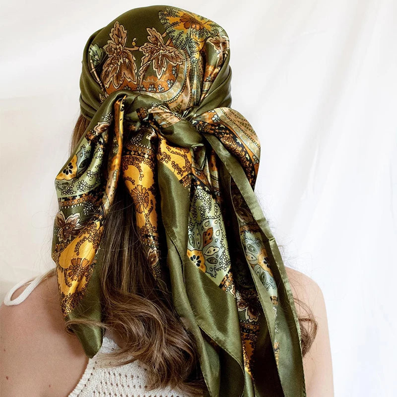 A close-up of a satin square scarf in olive green and gold floral print tied around a person's hair.