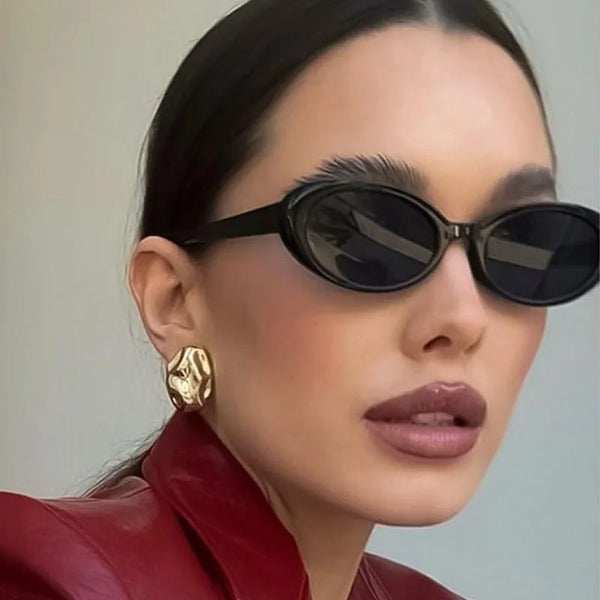 A close-up portrait of a woman wearing black retro oval sunglasses and gold earrings, with a red leather jacket visible.