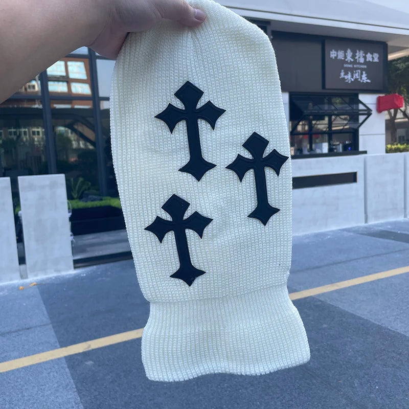 A white tactical knit beanie hat featuring five black cross symbols on the front, held by a hand outdoors.