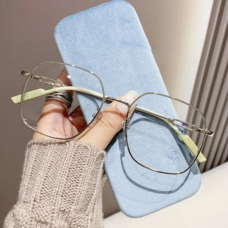 A pair of thin wire-frame reading glasses rests on a light blue cloth, held by a person wearing a beige knitted sweater.