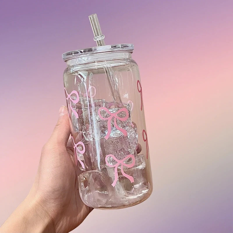 A hand holds a clear tumbler water bottle with pink bows and a straw against a light purple background.