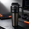 A matte black insulated stainless steel tumbler with a handle and straw sits on a dark desk next to a laptop.