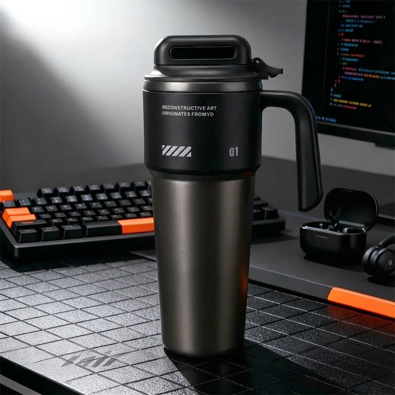 A matte black insulated stainless steel tumbler with a handle and straw sits on a dark desk next to a laptop.