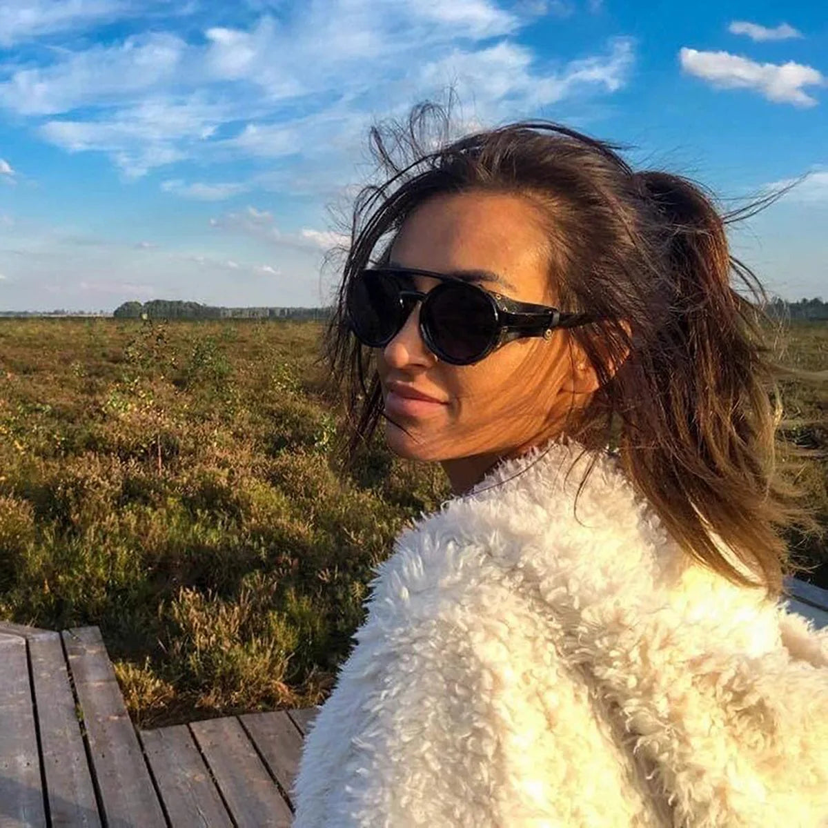A woman wearing black retro steampunk goggles sunglasses outdoors with a bright blue sky and green field background.