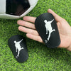 A hand holds a black mini shin guard for soccer, with a white silhouette of a player on it, placed on green grass.