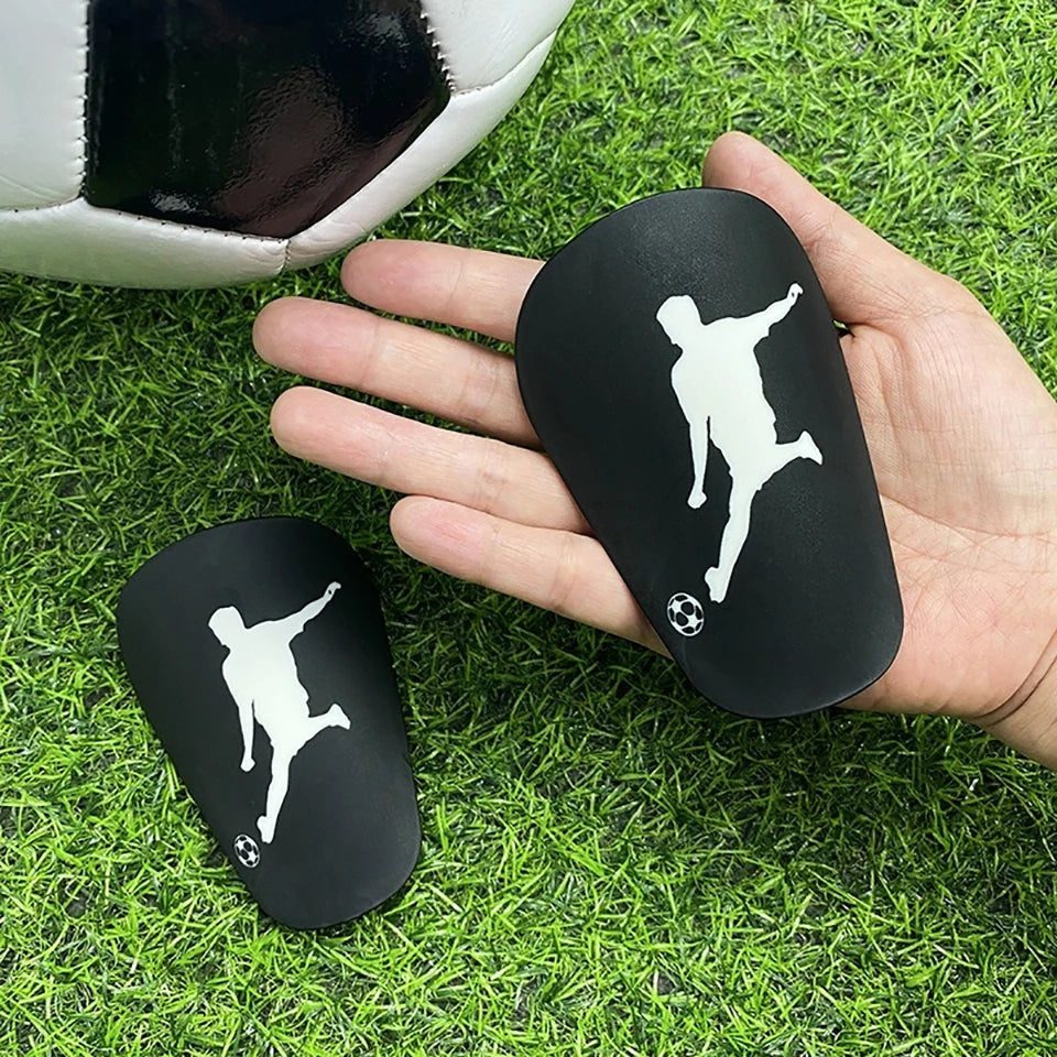 A hand holds a black mini shin guard for soccer, with a white silhouette of a player on it, placed on green grass.
