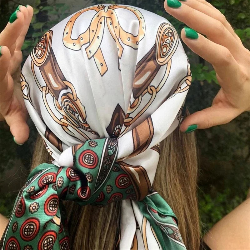 A hand ties a white satin square scarf with brown and green patterns into a bow for a hijab style.
