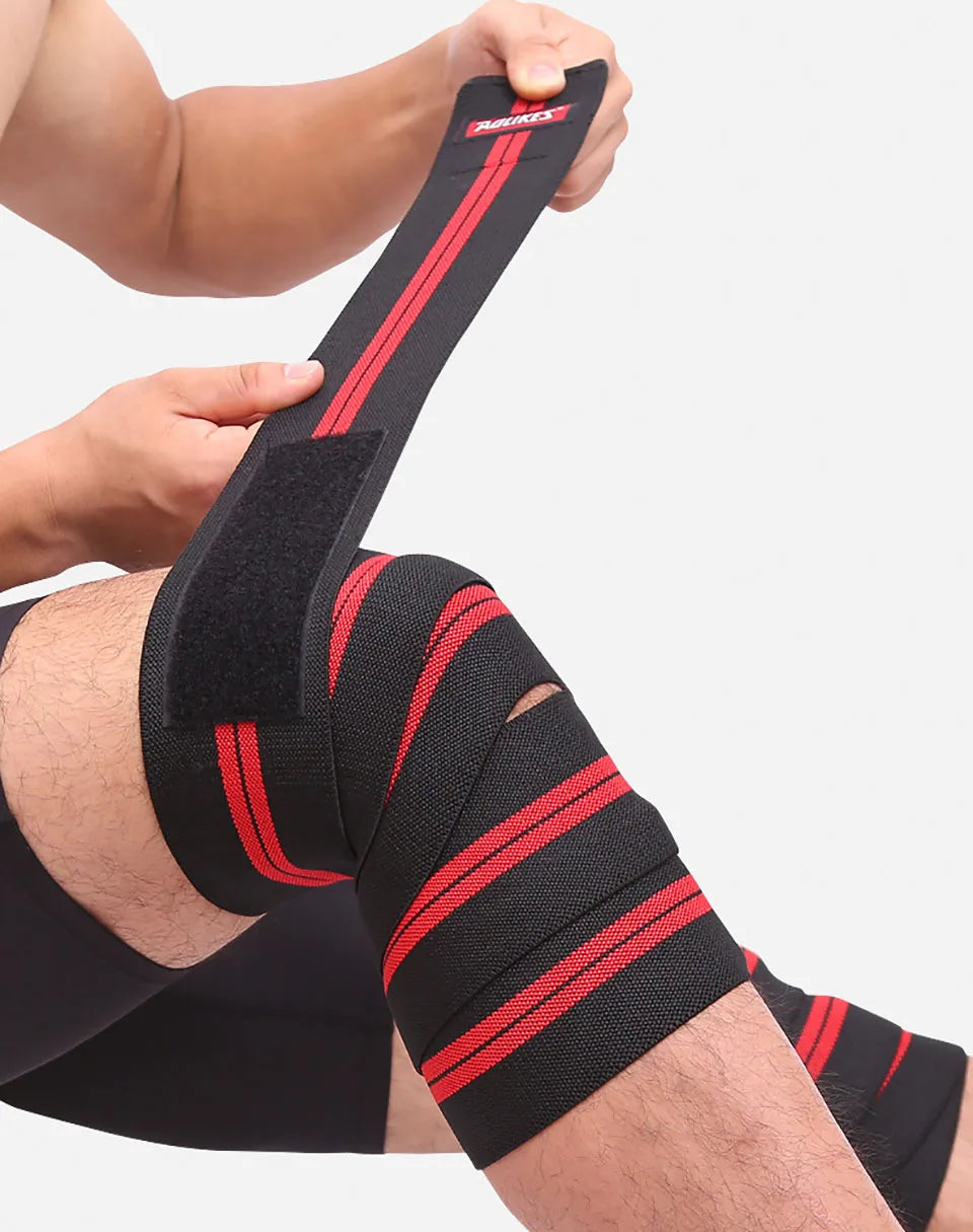 A person is applying black and red high-compression knee wraps to their leg for weightlifting.
