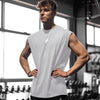 A muscular man wearing a white sleeveless fitness tank stands in a gym setting.