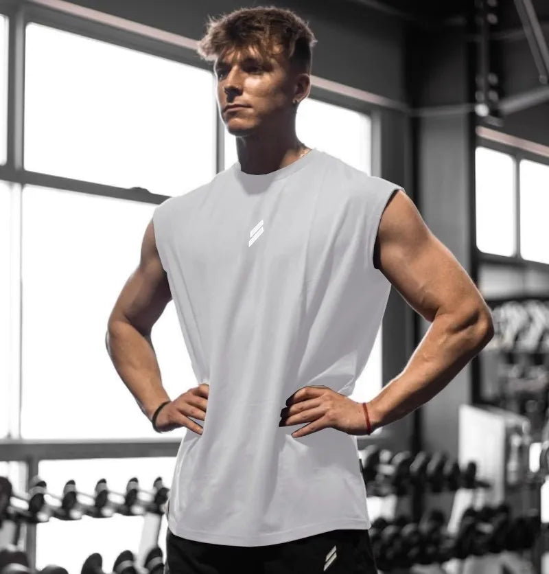 A muscular man wearing a white sleeveless fitness tank stands in a gym setting.