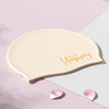 Gorra de silicona beige con la palabra Feliz escrita en dorado cursiva sobre un fondo rosa claro.