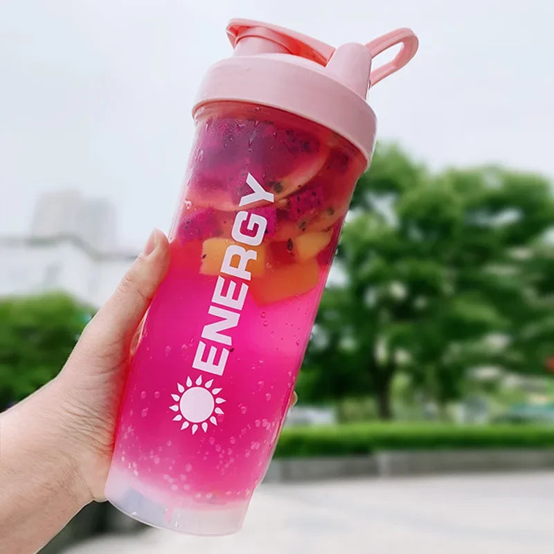 Eine Hand hält eine rosa, portable Protein-Shaker-Flasche mit leuchtend rosa Flüssigkeit und Fruchtstücken im Freien.