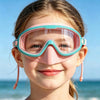 A smiling girl wearing pink and teal swimming goggles outdoors near the sea.