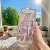 A hand holds a clear pink bow tumbler water bottle with a straw against a bright window background.