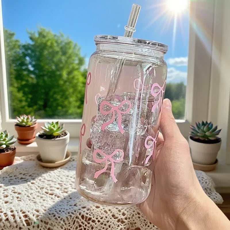 A hand holds a clear pink bow tumbler water bottle with a straw against a bright window background.