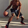 A muscular man wearing a black basketball shorts and a gold necklace is holding a basketball.