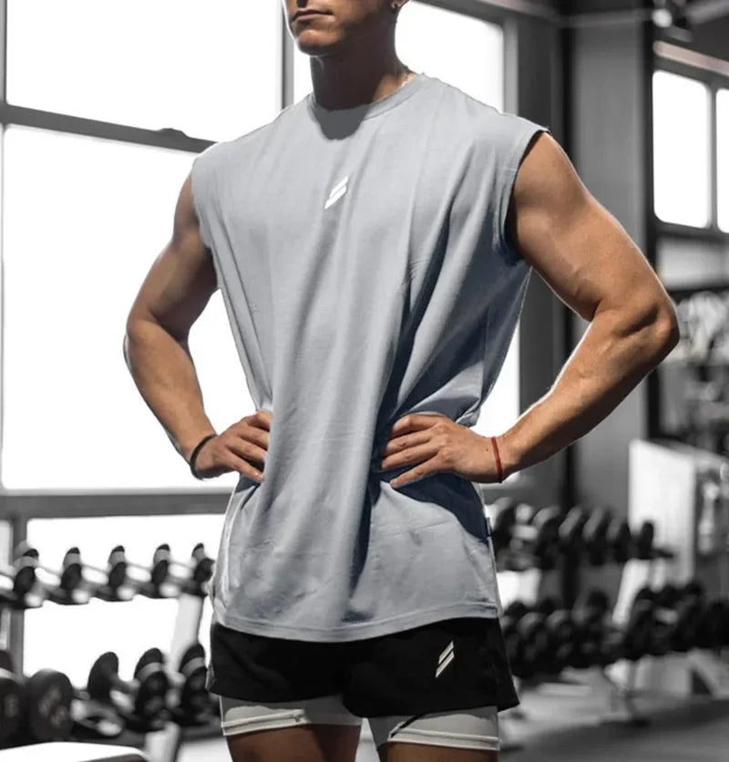 A muscular man wearing a light gray sleeveless fitness tank and black shorts in a gym setting.