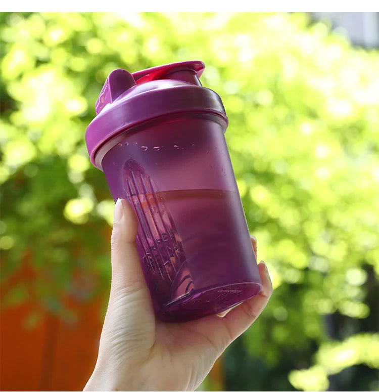 A hand holds a purple leak-proof sports shaker bottle against a bright green, leafy background.