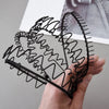 A hand holds a black metal hair band featuring intricate coiled and looped wire designs.