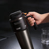 A hand holds a black insulated stainless steel tumbler with a handle and straw on a dark surface.
