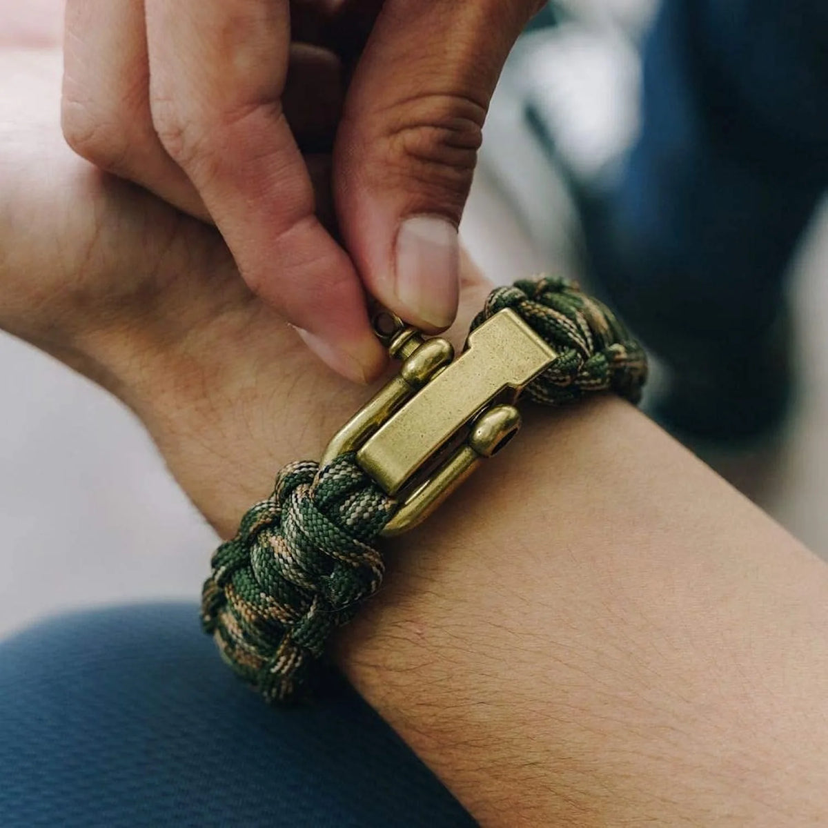 A hand is tying a tactical paracord survival bracelet with a bronze flag charm onto a wrist.