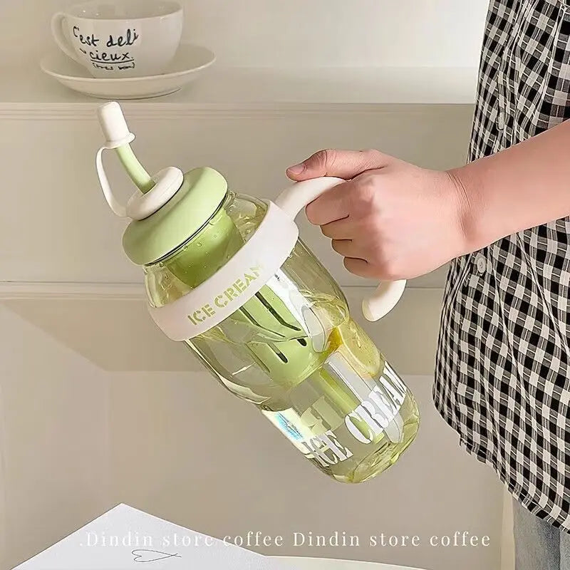 A hand holds a clear reusable water bottle with a straw and handle, filled with green liquid, against a white shelf.
