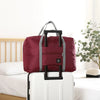 A maroon foldable travel duffel bag with a black handle is placed on a white rolling suitcase next to a wooden bed.