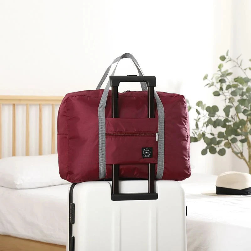 A maroon foldable travel duffel bag with a black handle is placed on a white rolling suitcase next to a wooden bed.