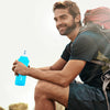A man wearing a dark shirt and backpack holds a bright blue silicone water bottle outdoors.