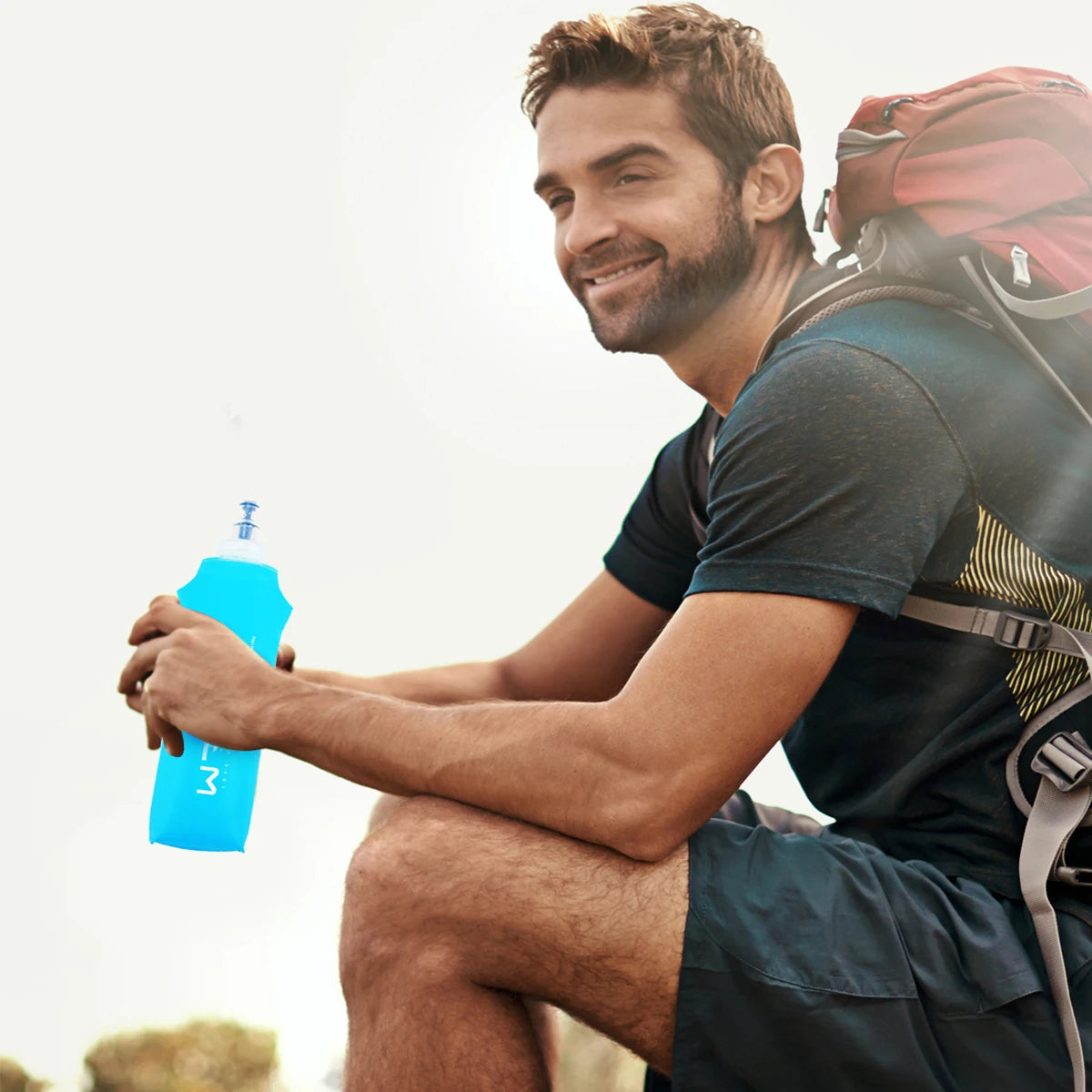 A man wearing a dark shirt and backpack holds a bright blue silicone water bottle outdoors.