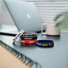 Primer plano de tres pulseras de silicona inspiradoras en negro, rojo y azul sobre una superficie oscura junto a una computad