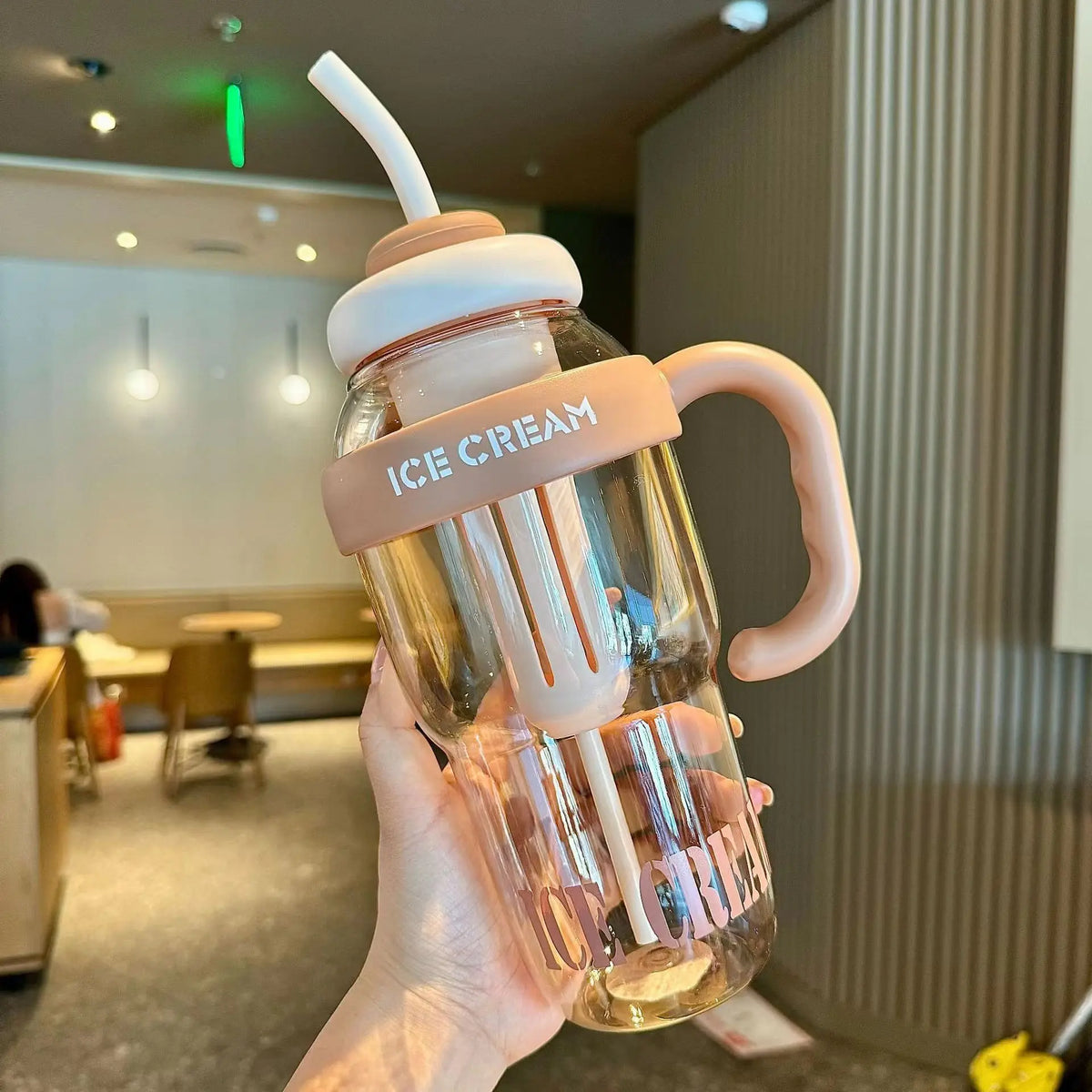 A hand holds a clear glass reusable water bottle with a straw and handle, labeled Ice Cream.