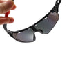 A hand is holding a pair of anti-glare sports sunglasses with dark lenses and black frames.