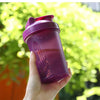A hand holds a purple leak-proof shaker bottle against a bright green, leafy background.