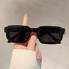 Une main tient des lunettes de soleil rectangulaires noires rétro sur un fond clair.