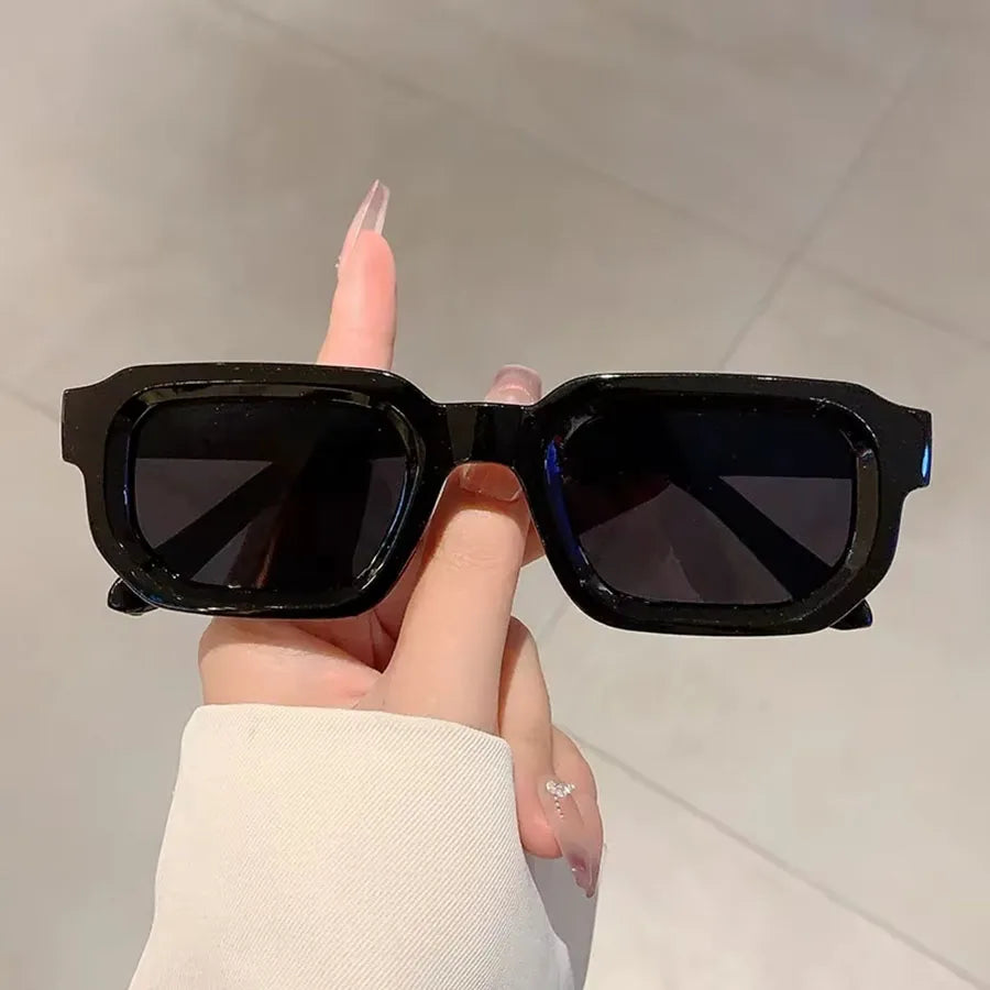 Une main tient des lunettes de soleil rectangulaires noires rétro sur un fond clair.