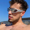 A man wearing silver-framed Y2K retro sports sunglasses outdoors against a blue sky.