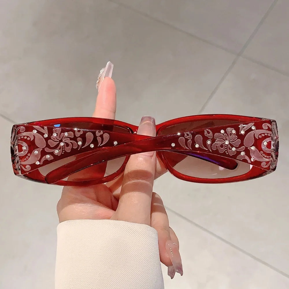 A hand holds a pair of red tinted Y2K cat eye rhinestone sunglasses.