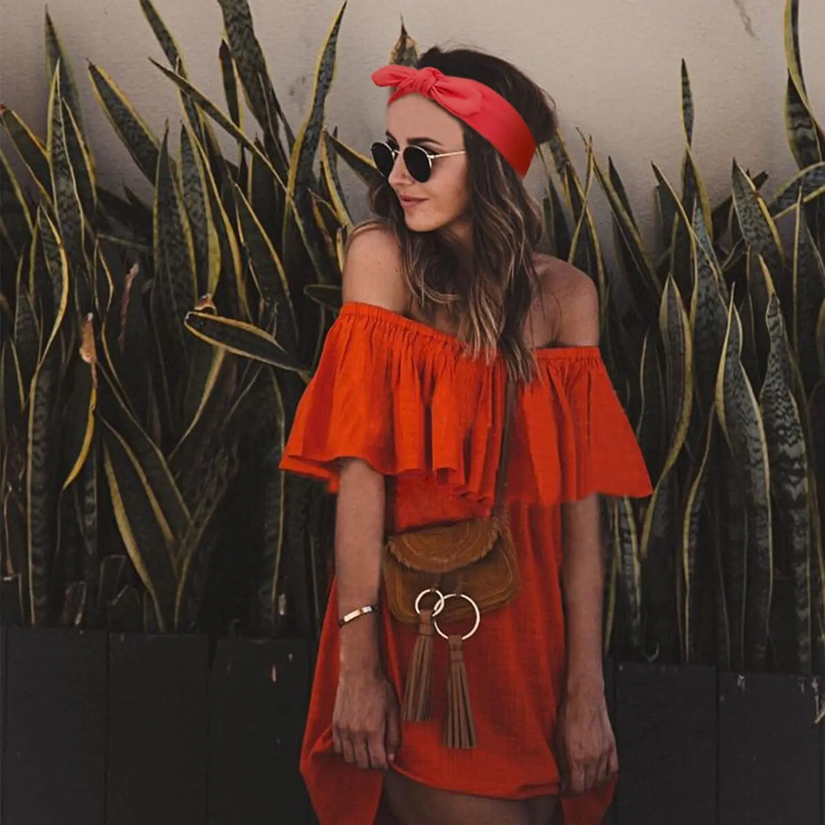 Eine Frau in einem orangefarbenen, schulterfreien Kleid und einem roten Stirnband steht vor grünen Sukkulentenpflanzen.