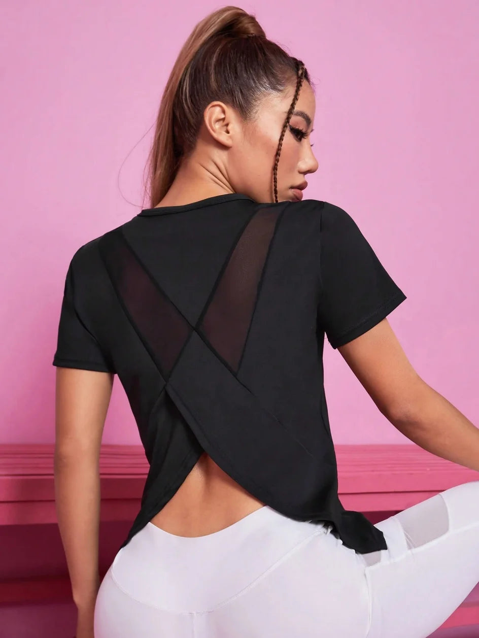 A woman wearing a black breathable summer yoga and fitness t-shirt with sheer paneling, posing against a pink background.
