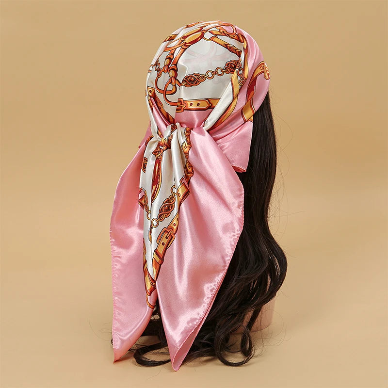 A pink satin square scarf with gold chain detailing is tied around a person's dark hair.