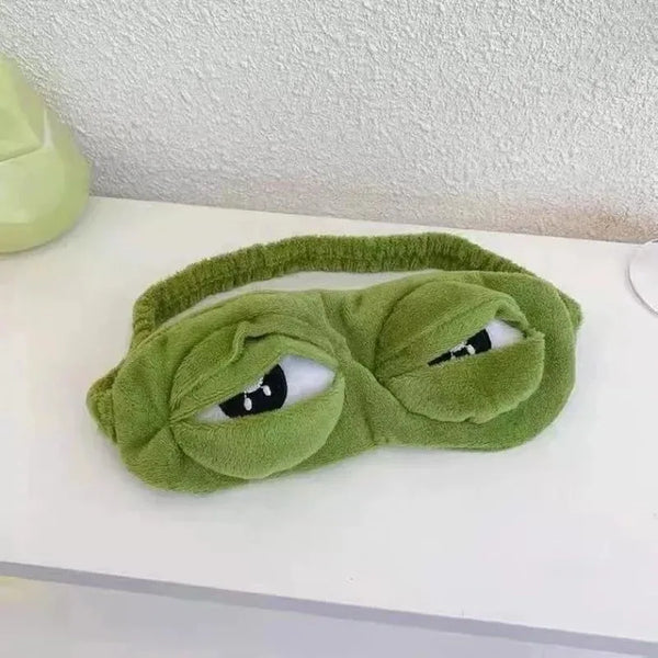 A green plush sleep eye mask shaped like a frog with closed eyes