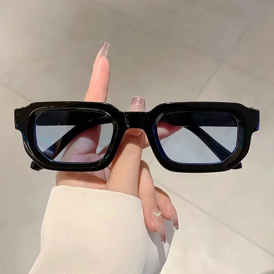 Une main tient des lunettes de soleil rectangulaires noires rétro sur un fond clair.