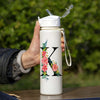 A hand holds a white insulated tumbler with a floral letter K design and a clear straw lid against a blurred green outdoor ba
