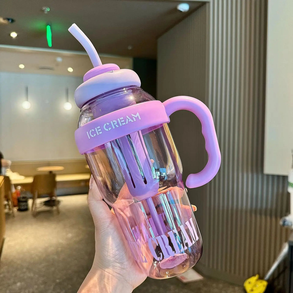 A hand holds a purple plastic sports water bottle with a pink lid and straw in an indoor setting.