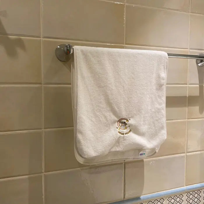 A white soft coral fleece face towel hanging on a metal bar against a tiled wall.