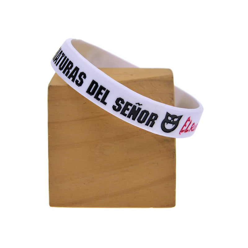 A white silicone wristband with black text reading MATURAS DEL SEÑOR and a small logo on a wooden block.